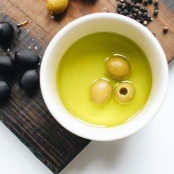 Weiße Schale mit grünem Olivenöl und 3 grünen Oliven auf dunklem Holzbrett mit schwarzen Oliven